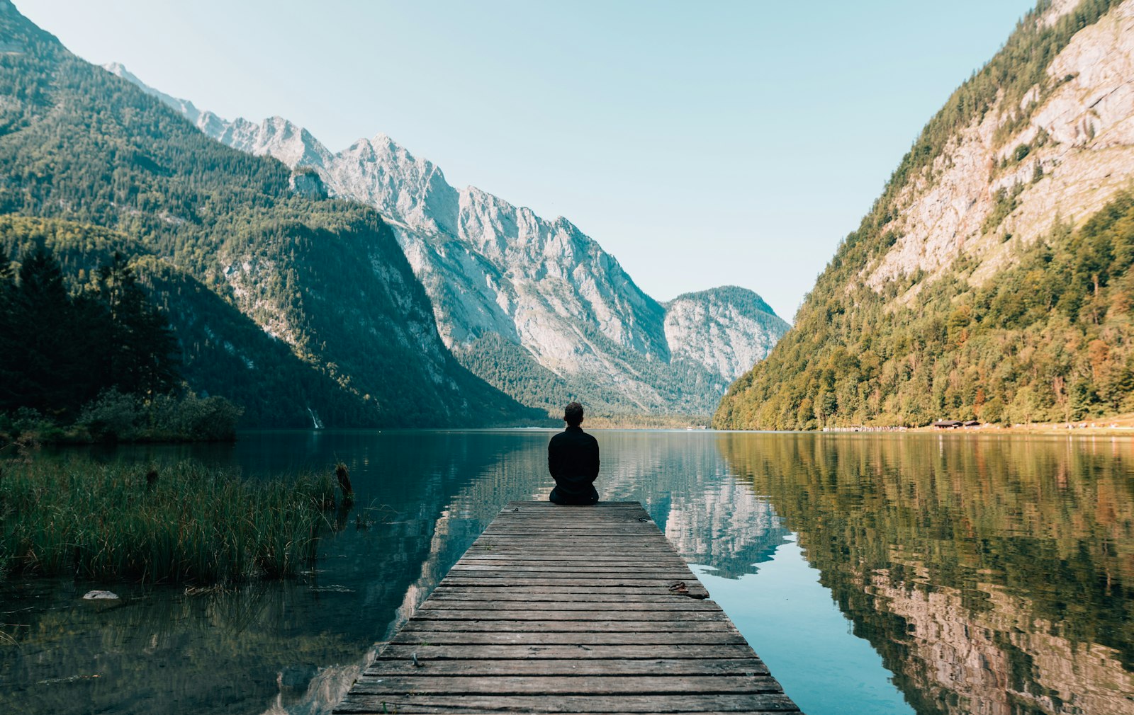 Persona meditando sobre un muelle de madera frente a un lago rodeado de montañas